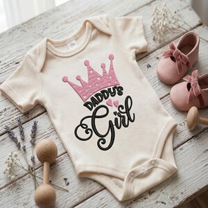 May include: A cream-colored short-sleeve baby bodysuit featuring an embroidered pink crown with small heart cutouts and the black embroidered text "DADDY'S Girl" with two small pink hearts positioned between the words on the chest area.
