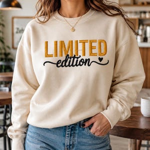 Pode incluir: Uma camisola de gola redonda cor de creme com a palavra LIMITED em letras maiúsculas bordadas a dourado e a palavra edition em letra cursiva preta, acompanhada por um pequeno símbolo de coração preto no lado direito do texto.