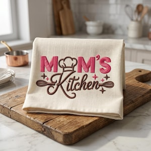 May include: A cream-colored kitchen towel featuring embroidered text that reads "MOM'S Kitchen" in pink and brown thread. The design includes a chef's hat icon, stylized whisk illustrations on the sides, and small star accents in pink and brown.