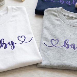 May include: White and gray t-shirts with the word "baby" embroidered in purple script, featuring heart accents. Additional folded shirts with the same design are visible in the background. A spool of purple thread and a measuring tape are in the background.
