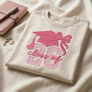 Peut inclure: Un t-shirt beige avec une broderie rose représentant une toque de diplômé avec un nœud et le texte "class of 26". Le motif comprend un grand nombre 26 dans une teinte rose clair avec un contour rose foncé, placé sous le graphique.