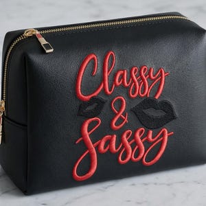 Può includere: Pochette per trucchi nera con cerniera dorata e la scritta ricamata "Classy & Sassy" in rosso. La borsa presenta due disegni di labbra nere. Prodotti per il trucco come rossetto, mascara e pennelli sono disposti intorno.