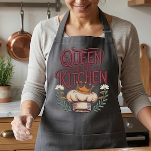 Puede incluir: Un delantal gris oscuro con el texto "Queen of the Kitchen" en una fuente decorativa, con un gorro de chef y detalles florales. El delantal se usa en un entorno de cocina con sartenes de cobre y una batidora de pie en el fondo.