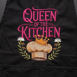 Op de afbeelding: Een zwart schort met geborduurde tekst Queen of the Kitchen in felroze. Onder de tekst staat een goudbruine koksmuts met een kleine kroon, geflankeerd door twee bloemstengels met witte bloemblaadjes en groene bladeren.