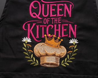 May include: A black apron featuring embroidered text that reads Queen of the Kitchen in bright pink. Below the text is a golden-brown chef hat topped with a small crown, flanked by two floral stems with white petals and green leaves.