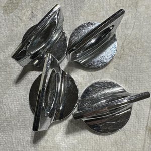 Può includere: Quattro manopole cromate lucide, con base circolare e maniglia triangolare. Le manopole sono disposte su una superficie strutturata grigio chiaro. Le maniglie sono angolate e hanno un design a coste.