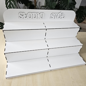 Pode incluir: Um expositor branco em camadas com as palavras "Słodki Stół" em letras brancas. O expositor tem três prateleiras e é feito de material branco com acabamento preto. O expositor está sobre uma superfície de madeira clara.