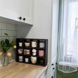 May include: A dark brown wooden shelf unit with twelve compartments, each holding a small decorative item. Items include miniature pots, cups, a radio, and a small statue. The shelf sits on a light wood countertop in a kitchen setting.