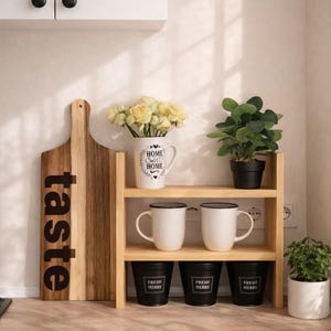 May include: A wooden cutting board with the word "taste" in black lettering. A wooden shelf holds white mugs, black containers labeled "Fresh Herbs", and a vase with flowers. A small potted plant is also visible.