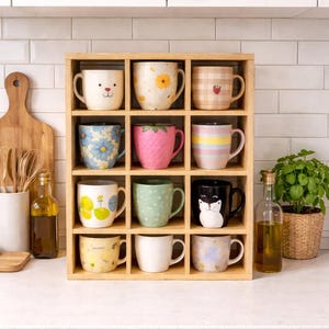 Puede incluir: Un estante de madera que muestra doce tazas de cerámica decorativas con varios diseños. Las tazas presentan patrones florales, caras de animales y otros gráficos coloridos. El estante está colocado contra un salpicadero y una encimera de azulejos blancos.