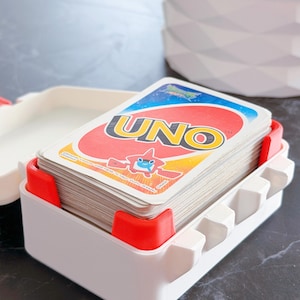 May include: A white and red UNO card holder with a deck of UNO cards. The holder has a rectangular shape with red corner supports and a series of white, hexagonal-shaped dividers. The UNO cards are visible, with the game's logo prominently displayed.