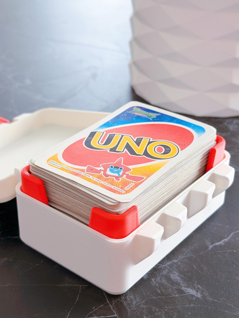 May include: A white and red UNO card holder with a deck of UNO cards. The holder has a rectangular shape with red corner supports and a series of white, hexagonal-shaped dividers. The UNO cards are visible, with the game's logo prominently displayed.