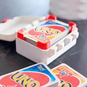May include: A white plastic UNO card holder with red accents, holding a deck of UNO cards. The cards have a red, yellow, blue, and green color scheme with the word "UNO" prominently displayed. Several cards are fanned out in front of the holder.