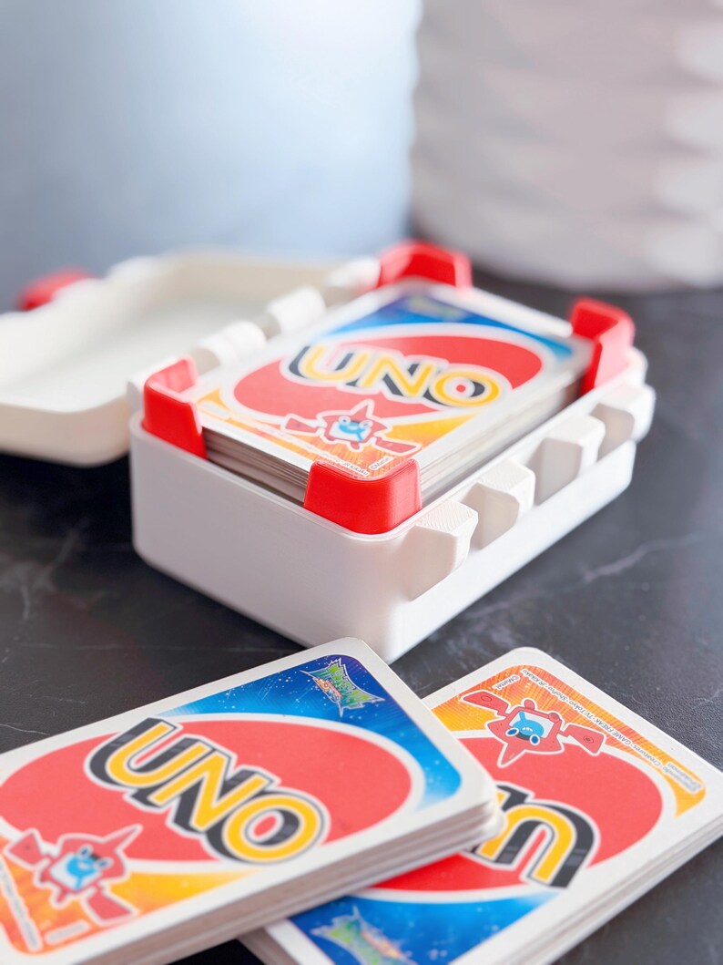 May include: A white plastic UNO card holder with red accents, holding a deck of UNO cards. The cards have a red, yellow, blue, and green color scheme with the word "UNO" prominently displayed. Several cards are fanned out in front of the holder.
