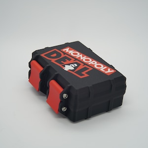 May include: Black and red Monopoly Deal card game case. The case has a black body with red accents and clasps. The Monopoly Deal logo is printed in red and white on the top of the case.