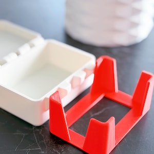 May include: A bright red, square-shaped object with four upright supports, likely a stand or holder. Beside it is a white, rectangular container with a hinged lid. The background includes a white, textured vase.