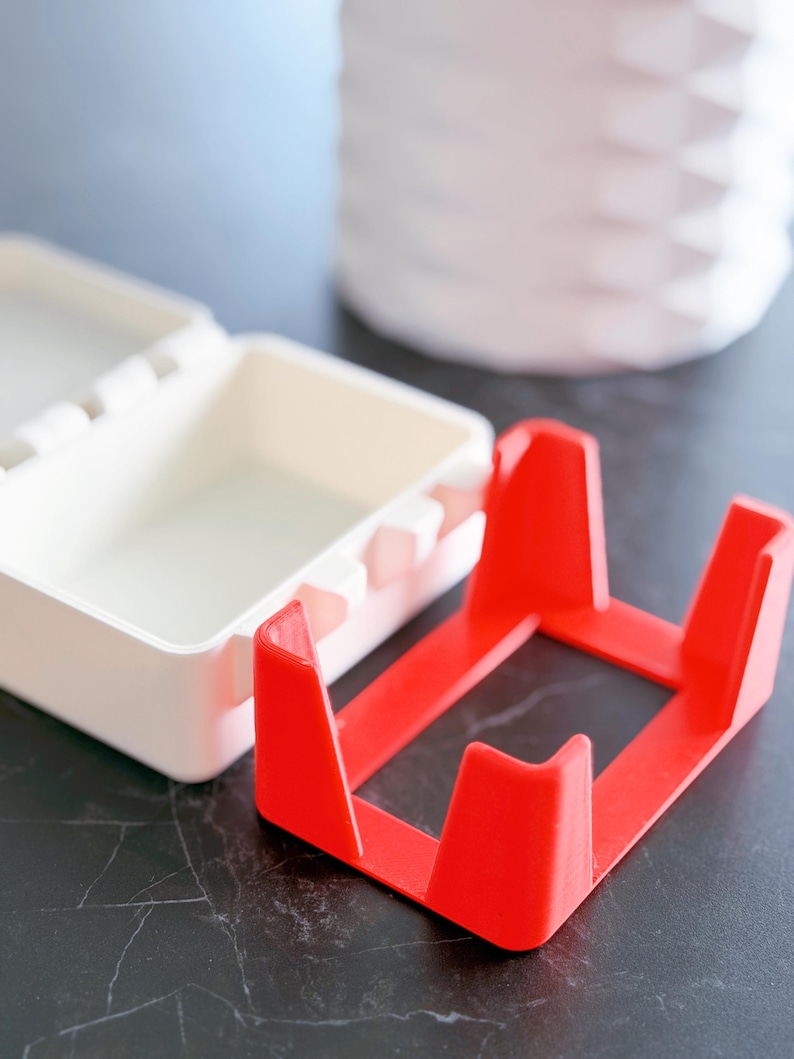 May include: A bright red, square-shaped object with four upright supports, likely a stand or holder. Beside it is a white, rectangular container with a hinged lid. The background includes a white, textured vase.