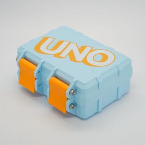May include: A light blue rectangular box with the word "UNO" in orange letters on the top. The box has orange clasps on the sides, designed to hold a card game.