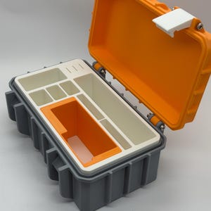 Peut inclure: Une boîte de rangement grise et orange ouverte, révélant un intérieur blanc avec des compartiments. Le couvercle orange est articulé et fixé par un loquet blanc. L'intérieur comporte des compartiments de différentes tailles.