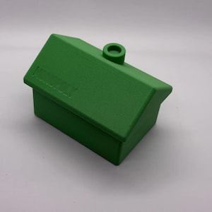 May include: A green plastic Monopoly house game piece featuring a gabled roof, a small chimney protrusion on the roof ridge, and the word Monopoly embossed in capital letters on the slanted roof surface, viewed from a high angle.