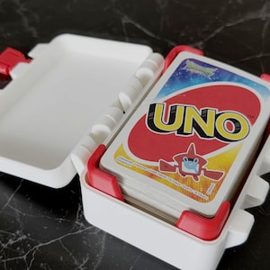 May include: A white plastic UNO card case with red accents, open to reveal a deck of UNO cards. The UNO cards have a red background with the word "UNO" in yellow and a cartoon character.