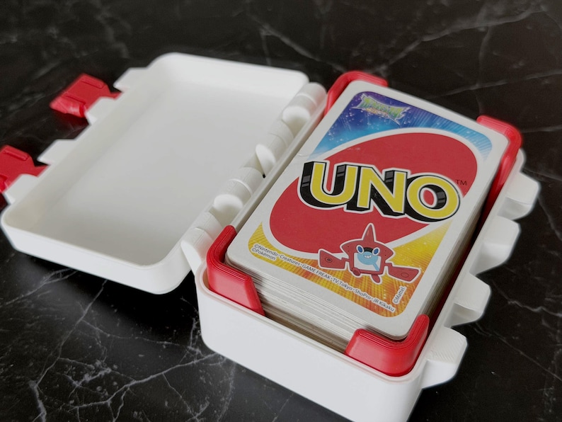 May include: A white plastic UNO card case with red accents, open to reveal a deck of UNO cards. The UNO cards have a red background with the word "UNO" in yellow and a cartoon character.