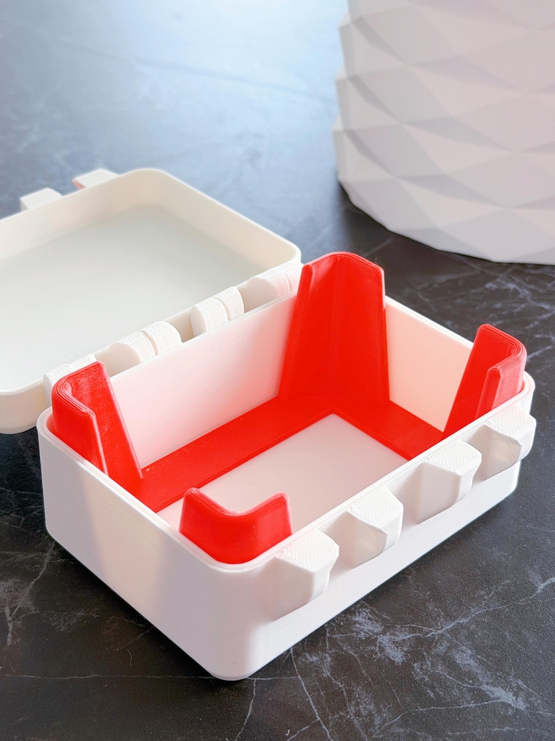 May include: A white and red plastic container with a lid. The container has a rectangular shape with red corner supports and a white interior. The lid is open, revealing the inside. The container is on a dark surface.