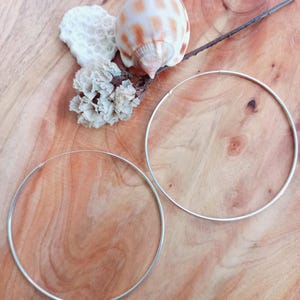 May include: Two silver hoop earrings sit on a wooden surface, with a seashell, dried flowers, and coral. The earrings are thin, circular, and have a simple design. The background is a warm wood grain. The earrings are approximately 7 cm in diameter.