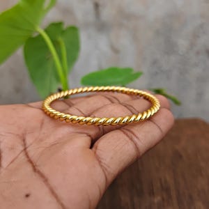 Pode incluir: Uma pulseira rígida de tom dourado polido com um design de corda torcida contínua. A banda metálica circular tem uma superfície lisa e reflexiva com um padrão espiral repetitivo que cria uma aparência texturizada em toda a circunferência.
