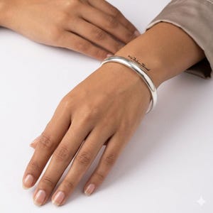 Puede incluir: Un brazalete de plata pulida con una superficie lisa y redondeada. El brazalete presenta un diseño minimalista y se lleva en la muñeca. El texto 925 Silver Thread está impreso en una fuente cursiva negra pequeña cerca del borde.