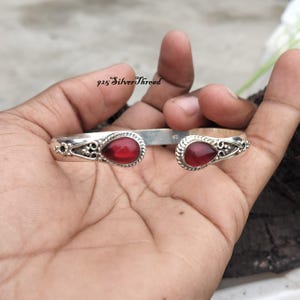 Peut inclure: Un bracelet manchette en argent avec deux pierres rouges en forme de goutte aux extrémités ouvertes. La bande métallique présente des détails en filigrane complexes autour des sertissages. Le texte 925 SilverThread est visible au-dessus.