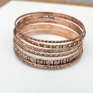 Peut inclure: Un empilement de cinq bracelets joncs en métal cuivré présentant divers motifs en relief, notamment des formes géométriques, des motifs floraux et des lignes texturées. Les bracelets sont disposés en grappe verticale.