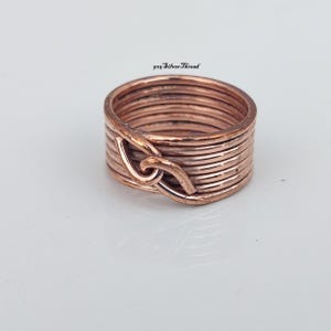 Op de afbeelding: Een koperkleurige ring opgebouwd uit meerdere gewikkelde draadlussen, met een centraal in elkaar grijpend knoopdetail. De tekst 925 SilverThread is zichtbaar boven de ring. Het ontwerp bestaat uit verschillende horizontale draadbanden.