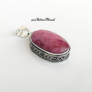 May include: An oval pendant featuring a faceted deep pink gemstone set in a sterling silver frame with intricate scrollwork and beaded edges. The metal bail is stamped with 925. The text 925 SilverThread is visible above the pendant.