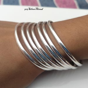 Peut inclure: Un ensemble de sept bracelets joncs en argent sterling 925 poli avec un profil lisse et arrondi. Les bracelets sont disposés en pile et présentent une finition métallique réfléchissante. Le texte 925 SilverThread est visible en haut.