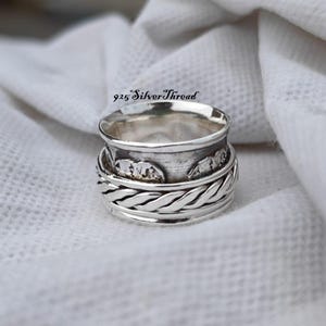 Peut inclure: Une bague rotative en argent sterling avec une bande centrale à la finition texturée et oxydée, bordée d'un motif tressé décoratif. La bague porte l'inscription 925 SilverThread et présente une surface métallique polie aux détails complexes.