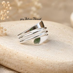 Puede incluir: Un anillo de plata pulida con un diseño envolvente de varias capas y extremos lisos y redondeados. La superficie metálica es brillante y reflectante. El texto 925 SilverThread es visible sobre el anillo en la imagen.