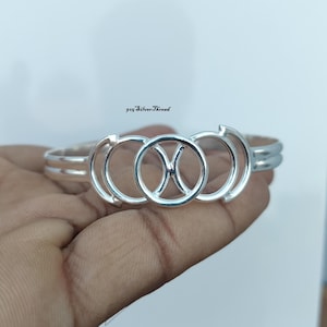 Könnte beinhalten: Ein poliertes Silberarmband mit einem zentralen kreisförmigen Design, das ein Fische-Sternzeichen-Symbol enthält und von zwei halbkreisförmigen Bögen flankiert wird. Das Metallband hat eine glatte, reflektierende Oberfläche. Der Text 925 SilverThread ist sichtbar.