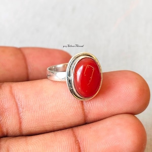 May include: A silver ring with an oval, bright red gemstone. The gemstone is set in a silver bezel with a gold-colored rim. The ring is held in a person's hand.