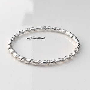 Può includere: Un sottile anello circolare in argento caratterizzato da una texture continua ritorta a forma di corda lungo tutta la fascia. Il testo 925 SilverThread è stampato in nero al centro dell'anello, indicando la composizione del materiale.