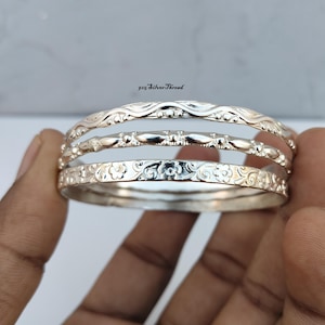May include: A set of three silver bangles featuring distinct textures. The top bangle has a wavy pattern, the middle has a repeating geometric design, and the bottom has an embossed floral motif. The text 925 SilverThread is visible above.
