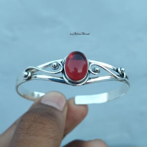 May include: A silver cuff bracelet featuring a central oval-shaped red gemstone. The metal band displays intricate swirling wirework patterns on both sides of the stone. The text 925 SilverThread is visible above the bracelet in black font.