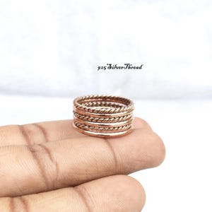 May include: A stack of five thin copper-colored rings featuring alternating smooth and twisted rope textures. The text 925 SilverThread is visible above the rings. The rings are metallic with a warm reddish-brown finish and a polished surface.