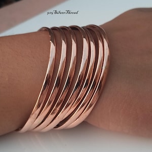 May include: A set of seven polished copper-coloured bangle bracelets with a smooth, reflective surface. The bracelets are stacked together, creating a wide band appearance. The text 925 SilverThread is visible in the upper portion of the image.