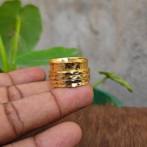 Peut inclure: Une bague en métal doré présentant une large bande avec une texture martelée et alvéolée. Le design se compose d'une bande centrale flanquée de deux bandes plus fines, partageant toutes la même finition métallique réfléchissante et irrégulière.