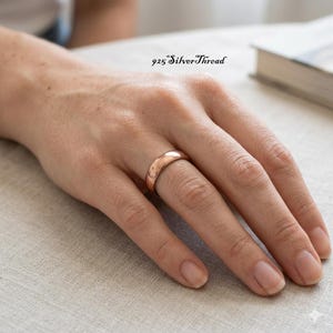 Könnte beinhalten: Ein glatter, polierter Bandring in Roségold-Optik am Ringfinger. Der Text 925 SilverThread ist in schwarzer Schreibschrift über der Hand zu sehen. Der Ring hat ein schlichtes, abgerundetes Profil mit einer reflektierenden metallischen Oberfläche.