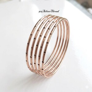 Peut inclure: Un bracelet jonc de couleur or rose composé de sept anneaux circulaires fins et interconnectés. Chaque anneau présente une série de petites indentations verticales régulièrement espacées. Le texte 925 SilverThread est visible.