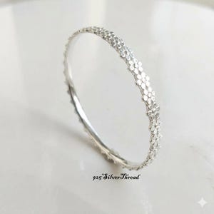 Puede incluir: Una pulsera de plata fina con un patrón continuo de pequeñas formas de flores en relieve a lo largo de la banda exterior. El metal tiene un acabado brillante y reflectante. El texto 925 SilverThread está impreso en negro en la parte inferior.