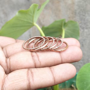 Puede incluir: Un conjunto de cinco anillos metálicos finos en tono cobre. La colección incluye tres anillos con una textura trenzada similar a una cuerda y dos anillos con una superficie lisa y sencilla. Los anillos están dispuestos en fila, mostrando su acabado metálico.