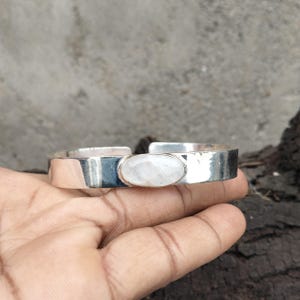 Könnte beinhalten: Ein silberner Armreif mit glatter, polierter Oberfläche und offenem Design. In der Mitte des Armreifs befindet sich ein einzelner, ovaler, durchscheinender weißer Mondstein-Cabochon, der in einer einfachen, klaren Metallfassung gehalten wird.